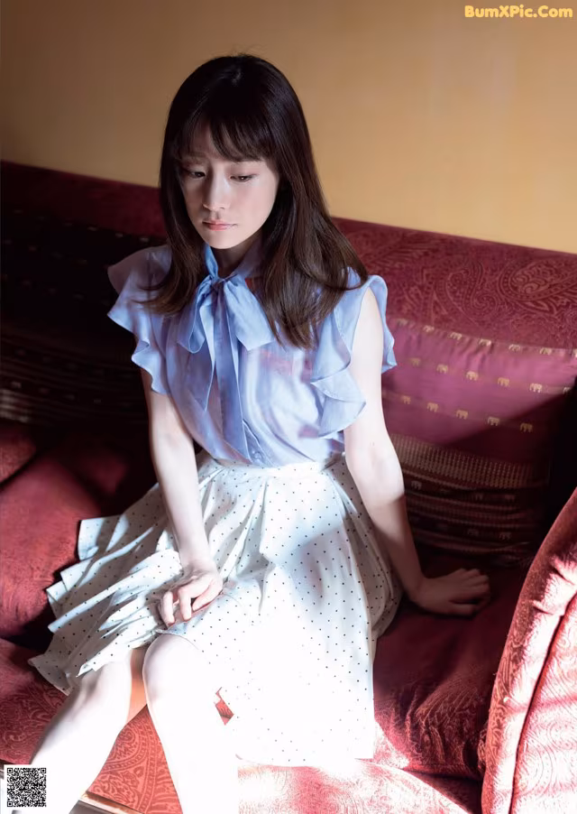 A woman sitting on a red couch wearing a blue shirt and white skirt.